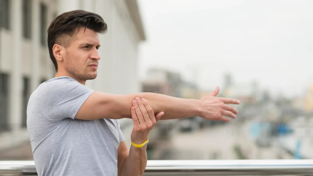 Hombre joven con camiseta gris y pulsera amarilla estirando su brazo derecho al aire libre, con un fondo urbano difuminado.